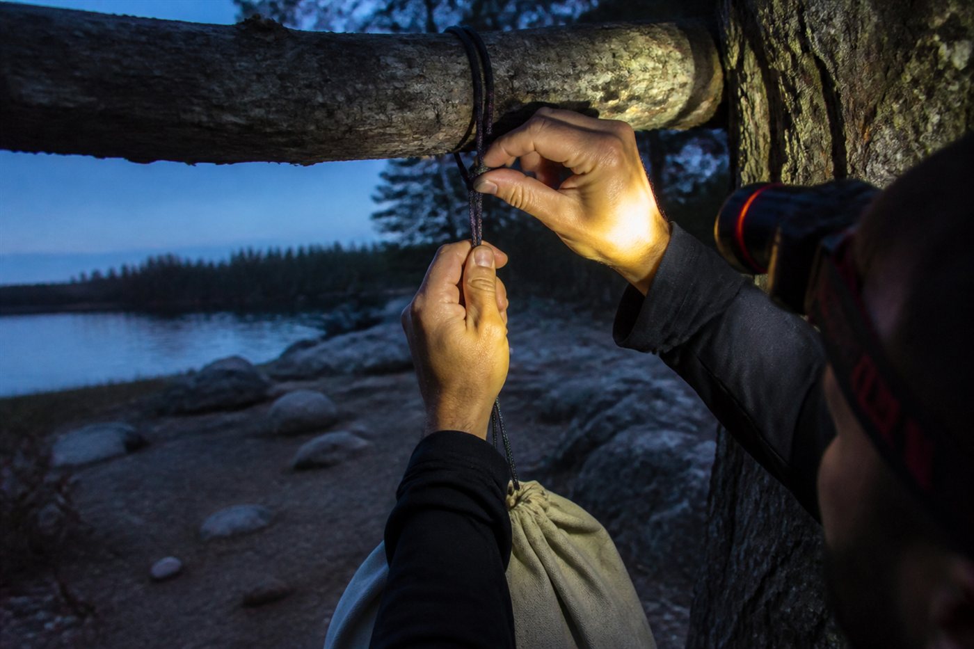 Hands tying a food-hang cord by headlamp at dusk